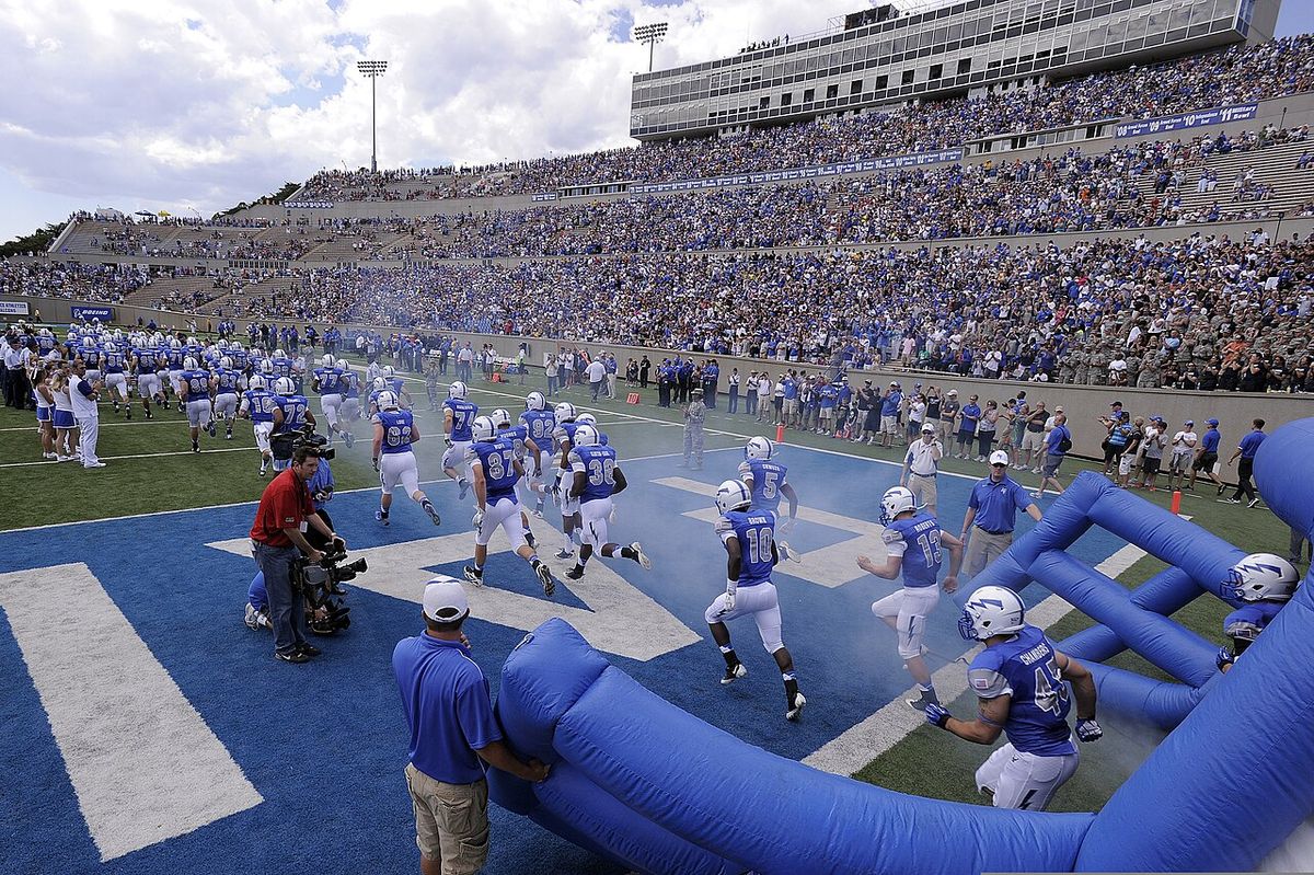 Air Force football players in blue run onto the field through an inflatable tunnel in front of a big crowd.