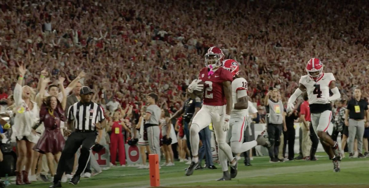 An Alabama receiver in a crimson jersey runs into the end zone with the ball as Georgia defenders in white chase him and the packed crowd behind them celebrates.