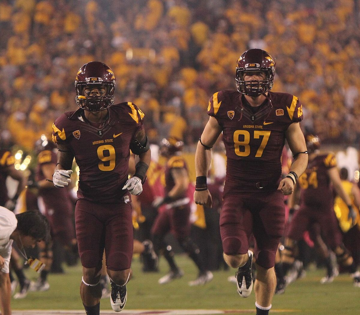 Two Arizona State players in maroon uniforms jog onto the field under the lights.