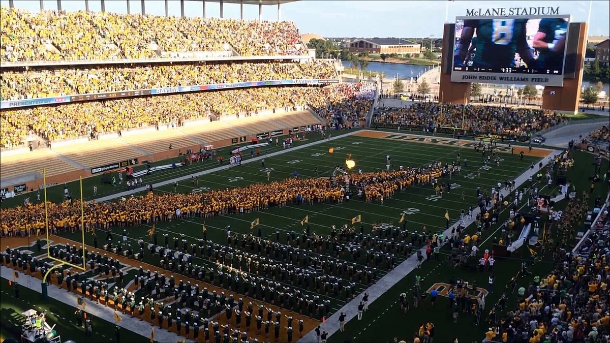 A packed stadium in green and gold, with a huge crowd of students in gold shirts forming a tunnel across the field next to the marching band, under a video board reading McLane Stadium.