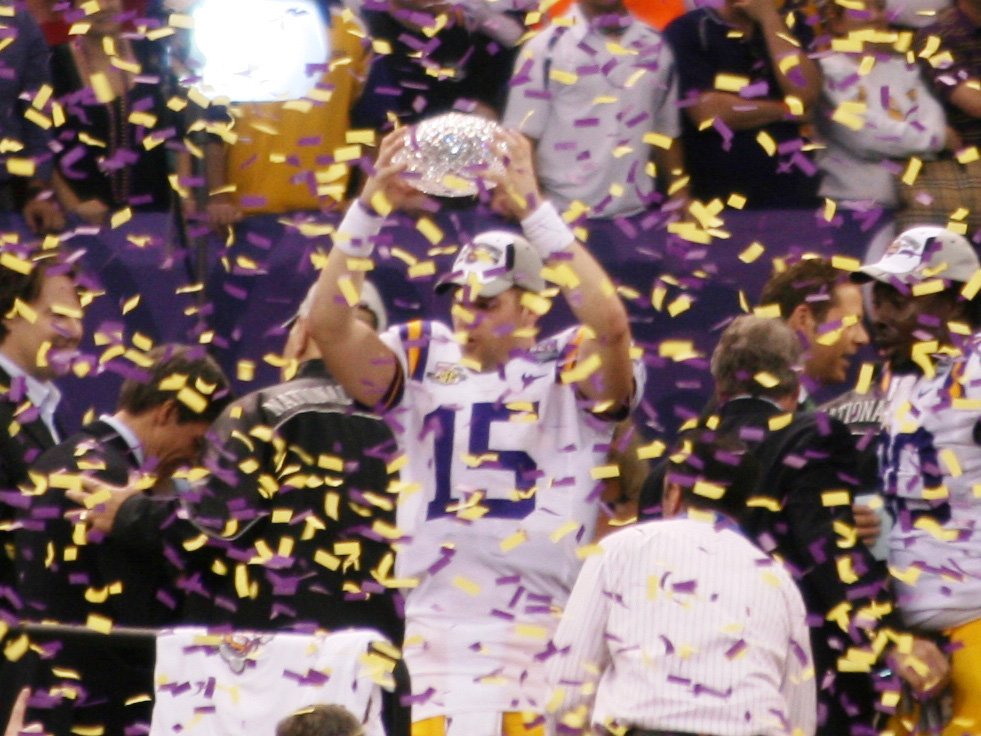 A football player in a white LSU jersey, number 15, lifts a crystal football trophy over his head as purple and gold confetti falls around him.