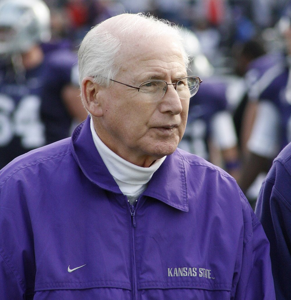 Kansas State coach Bill Snyder, with white hair and glasses, wearing a purple Kansas State jacket on the sideline, with players in purple behind him.