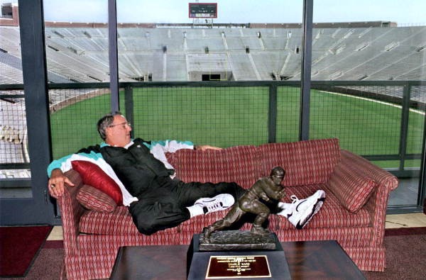 Bobby Bowden stretched out on a couch in a stadium suite, with a Heisman Trophy on the table in front of him and an empty stadium behind the glass.
