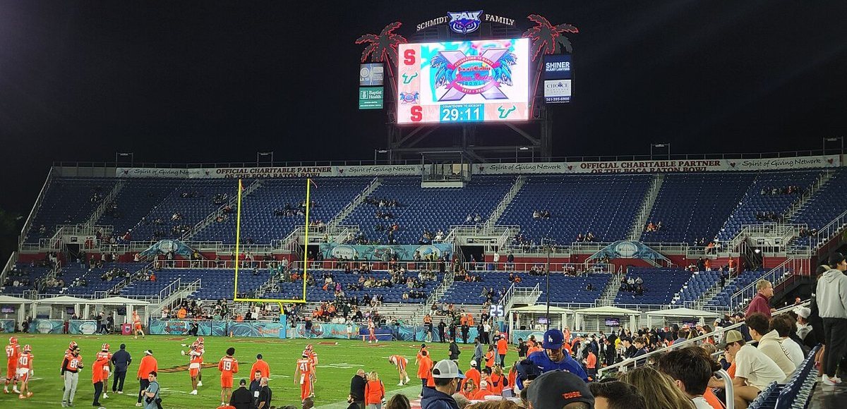 A stadium at night with a large video scoreboard counting down to kickoff, players warming up on the field below.