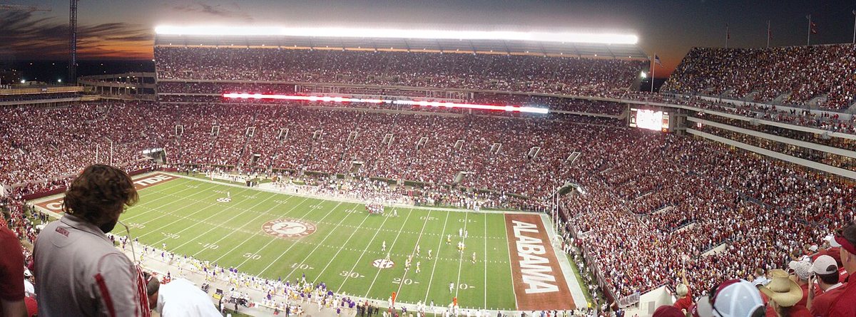 A wide view of a packed Bryant-Denny Stadium under the lights at dusk, with the crimson ALABAMA end zone in the foreground and the sky turning orange behind the upper deck.