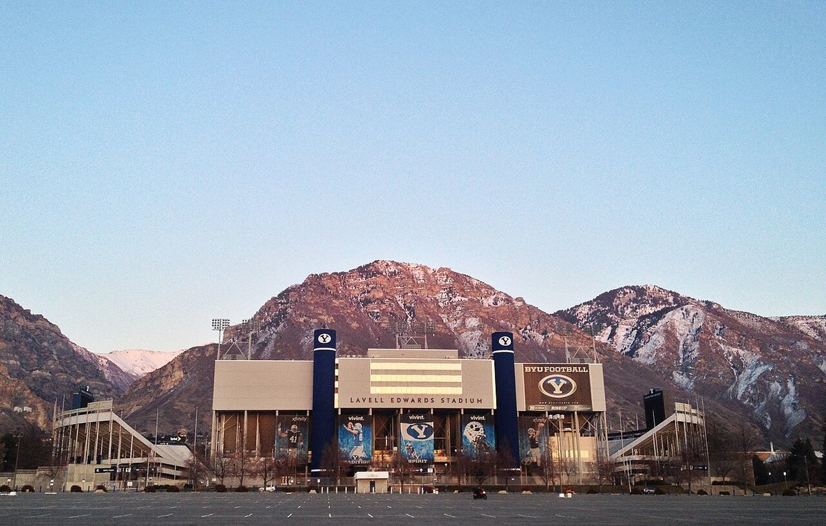 A large football stadium with two tall blue towers and a sign reading LaVell Edwards Stadium, in front of snow-dusted mountains at dusk, with an empty parking lot in the foreground.