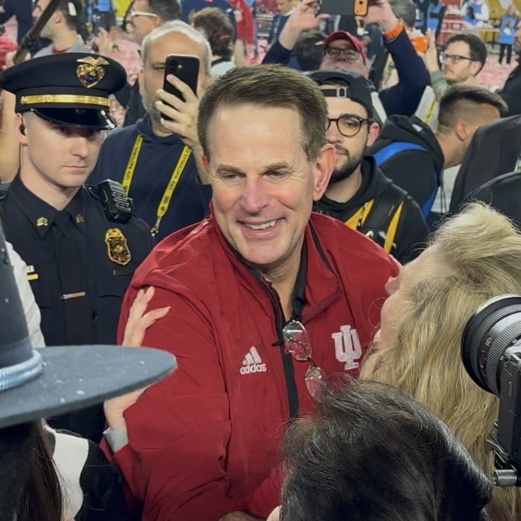 Indiana coach Curt Cignetti, in a red Indiana jacket, smiles as people crowd around him on the field after the national championship game.