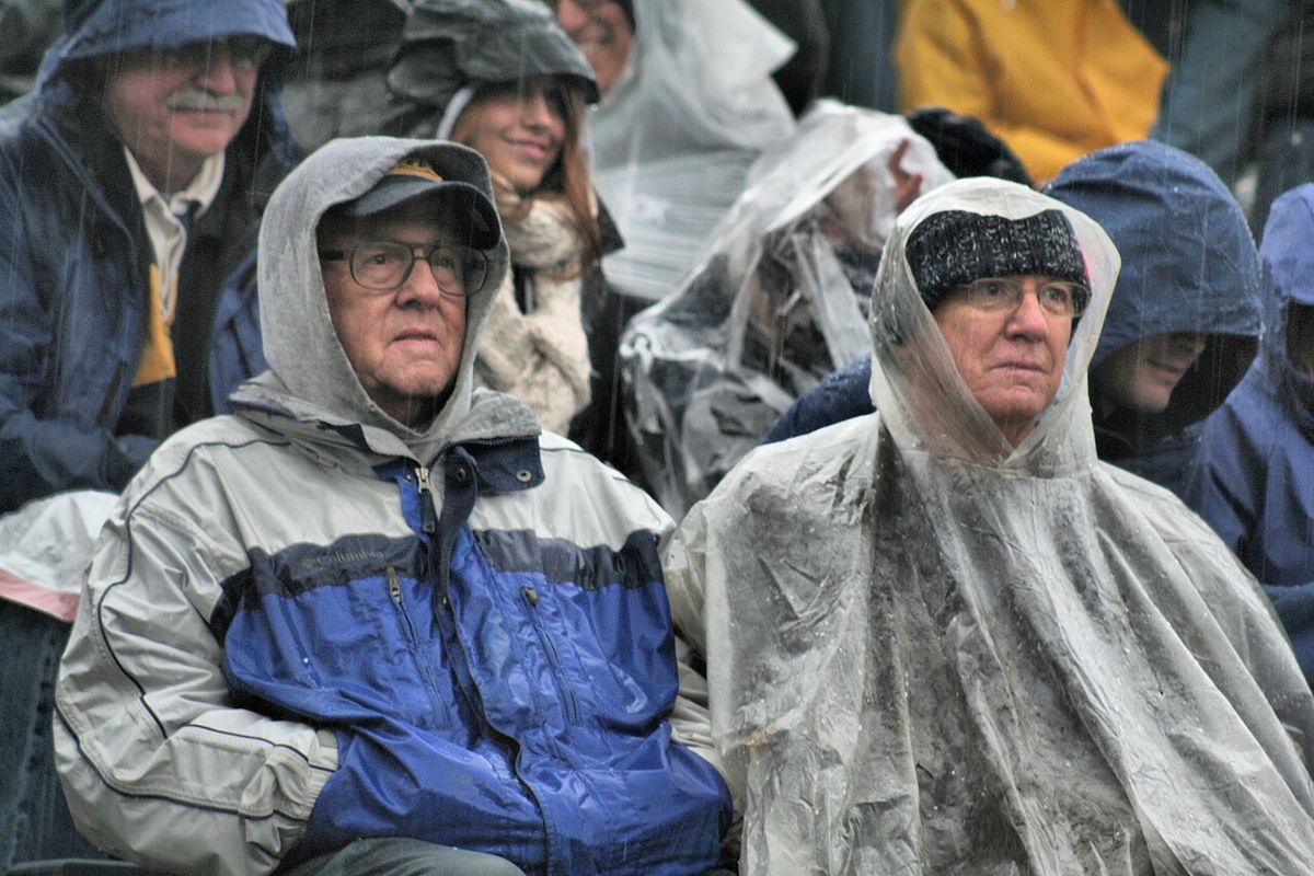 Two older football fans bundled in rain jackets and a clear plastic poncho, sitting in the rain.