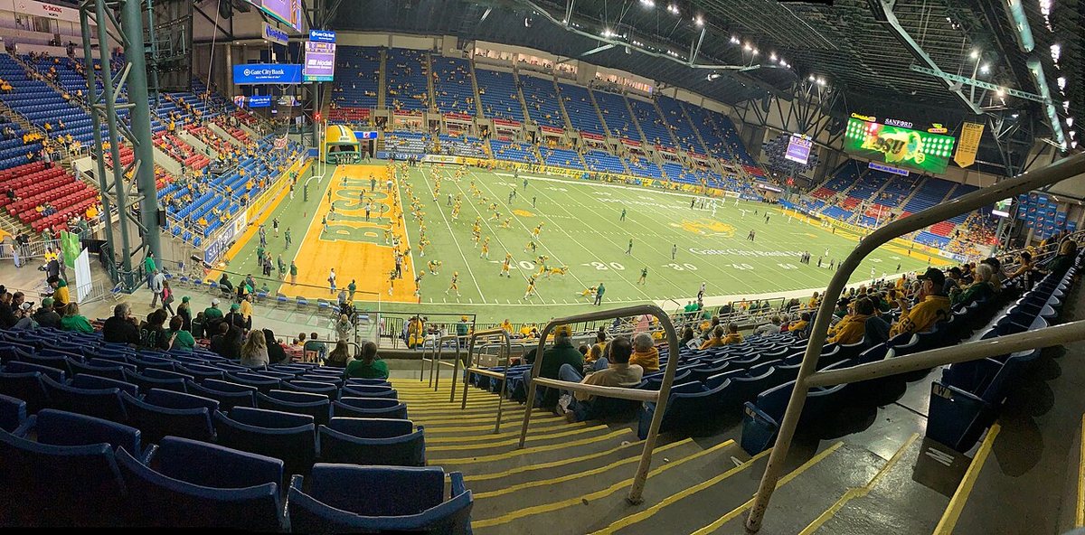 Inside a domed stadium with blue seats and a green field with a yellow end zone reading BISON, as North Dakota State players warm up before a game.