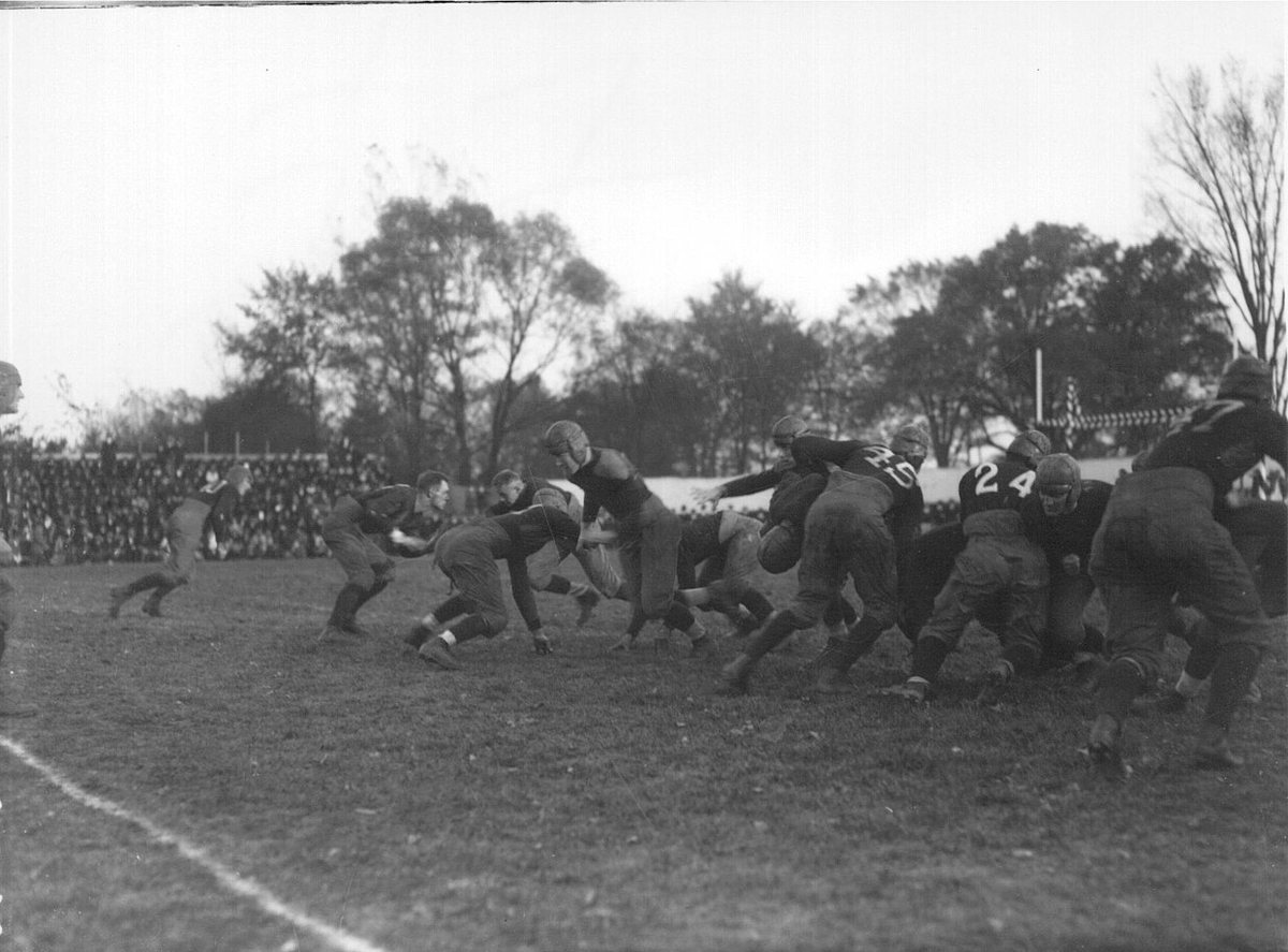 A black-and-white photo of a crowd of football players in leather helmets piled up near the goal line, with spectators and trees in the background.