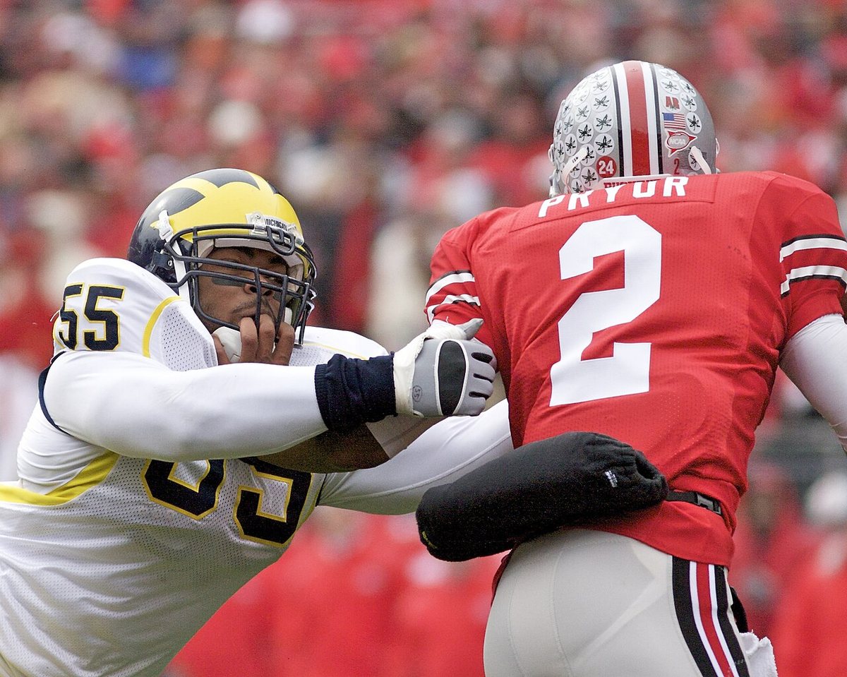 A Michigan defender in a white jersey with a maize helmet reaches out to grab Ohio State's quarterback, number 2 in red, from behind.