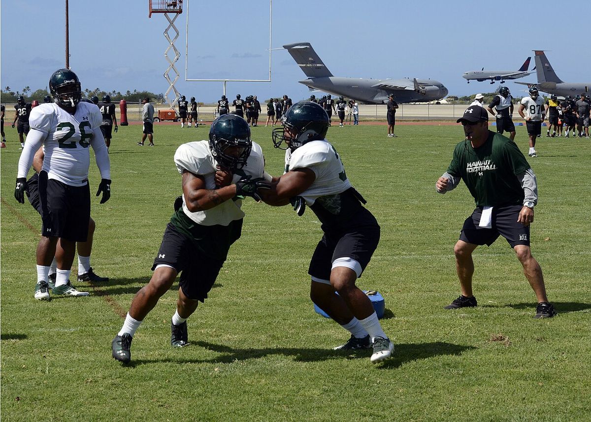 Hawai'i football players in practice gear run drills on a grass field, with military cargo planes parked in the background.