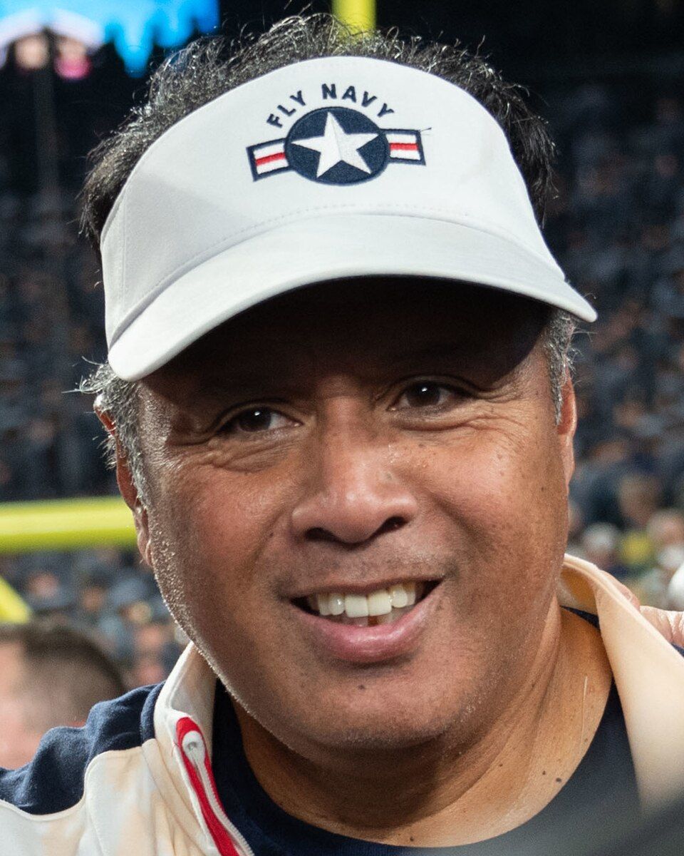 Navy head coach Ken Niumatalolo smiling on the field, wearing a white Fly Navy visor.