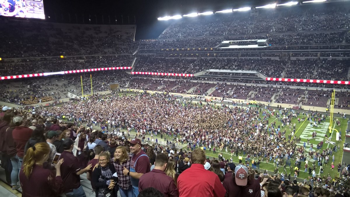 Thousands of fans in maroon flood the field at a packed, brightly lit Kyle Field at night, while fans in the stands celebrate in the foreground.