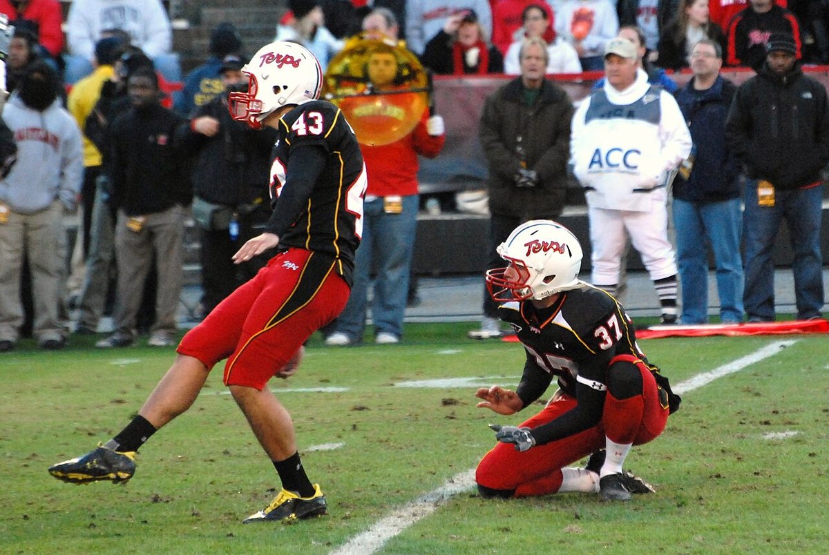 A Maryland kicker in a red and black uniform follows through after a kick while a kneeling teammate holds the spot, with fans and an ACC official in the background.