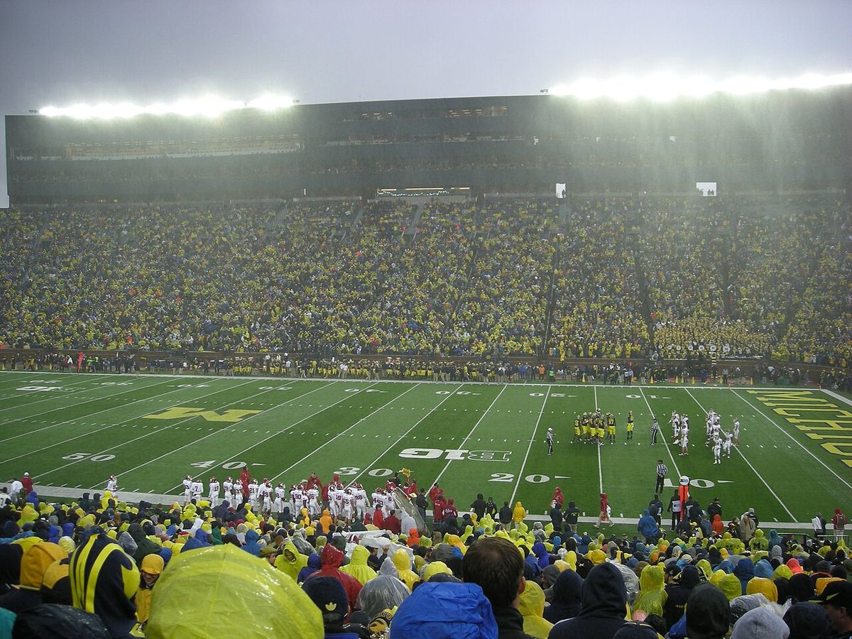 Michigan Stadium in heavy rain, with fans in yellow and blue ponchos in the foreground.