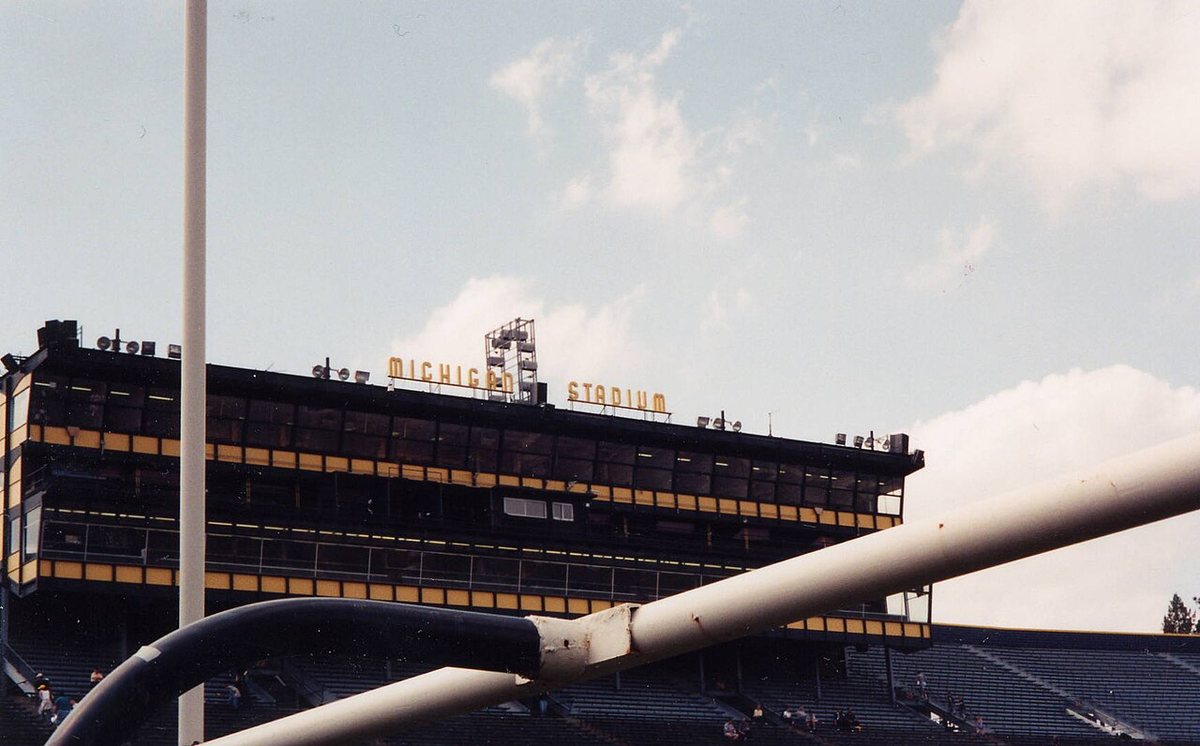A tall press box with dark windows and maize trim and the words Michigan Stadium on top, seen through white goal posts on a cloudy day.