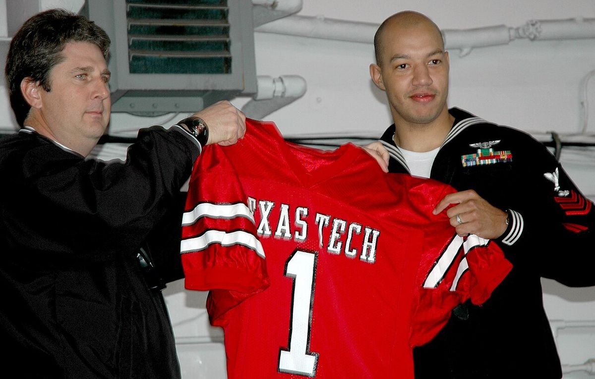 Texas Tech coach Mike Leach holds up a red Texas Tech number 1 jersey with a Navy sailor in dress blues.