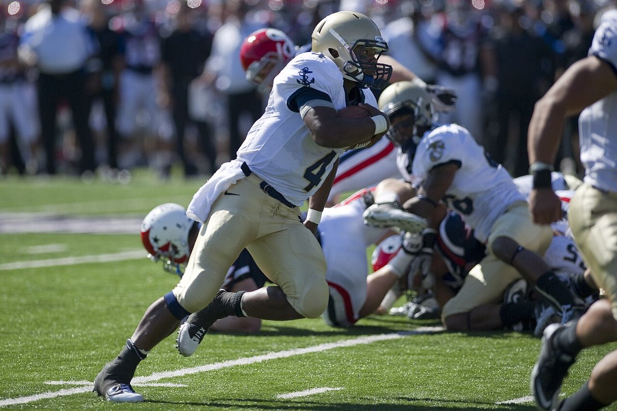 A Navy ball carrier in a white jersey and gold pants runs with the football past a pile of players, with Air Force players in white and red helmets on the ground.