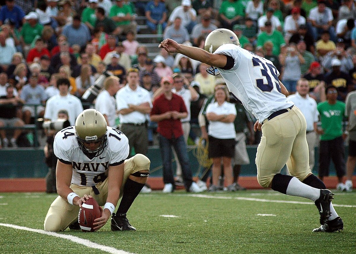 A Navy kicker in a white jersey and gold pants follows through on a kick while a teammate in a gold helmet holds the ball, with fans in green in the stands behind them.