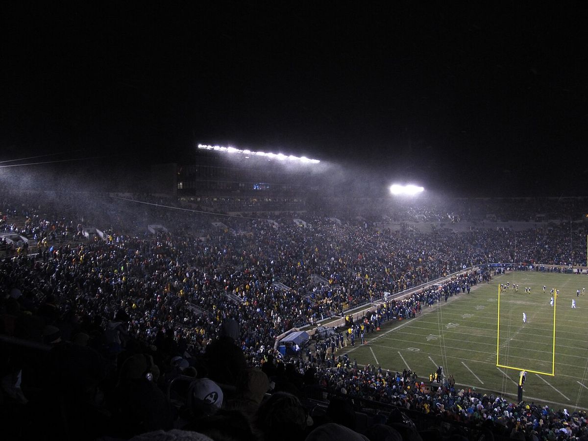 Snow blowing through the lights over a packed Notre Dame Stadium at night.