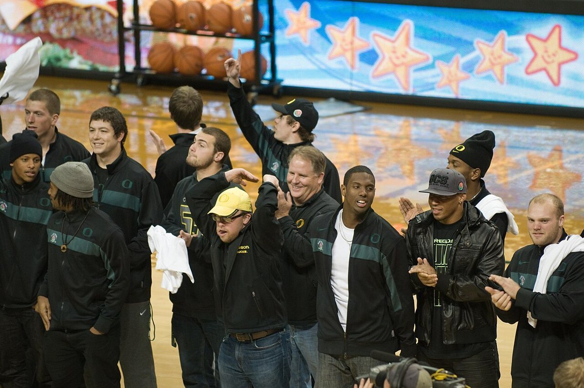 A group of Oregon football players in dark team jackets and caps cheer and wave towels on a basketball court.