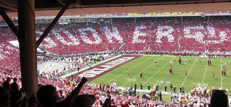 A packed stadium seen from under the upper deck, with fans in red and white holding up cards that spell out giant letters across the stands, and Oklahoma written in the end zone.