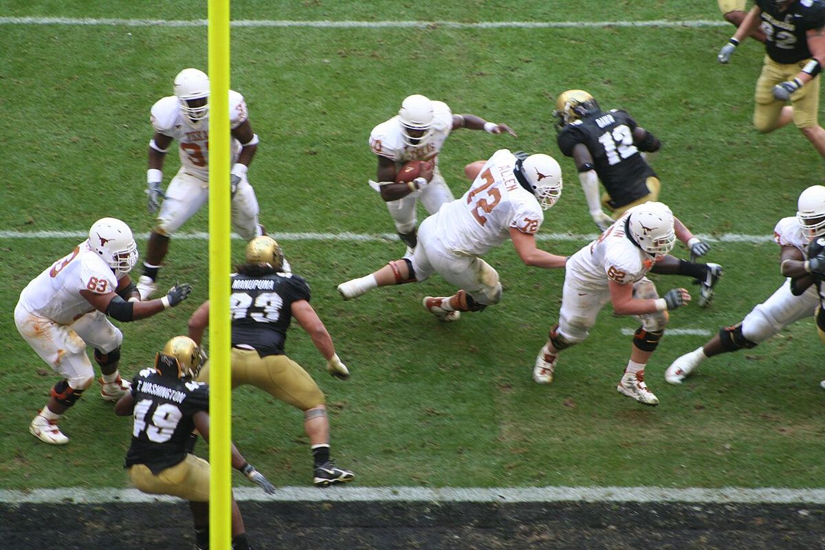 Texas players in white uniforms run through Colorado defenders in black jerseys near the goal line.