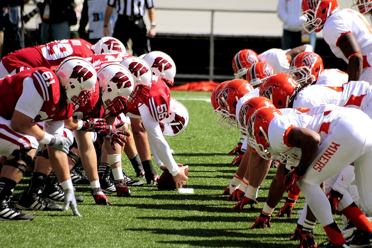 Looking down the line of scrimmage: Wisconsin linemen in red and white crouch facing UTEP linemen in white and orange, a moment before the snap.